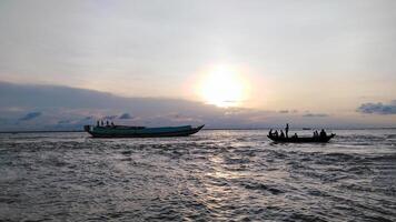 A serene sunset over a body of water. Two boats, one larger and one smaller, are visible. The partly cloudy sky and warm glow create a tranquil atmosphere. photo