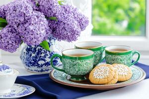 Elegant vintage tea set with green cups, cookies, and lilac flowers on a navy tablecloth in a bright setting photo