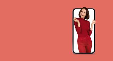 A person stands beside a large smartphone screen, smiling and animated while engaging with a digital application. The setting features a vibrant coral background, emphasizing the connection. photo