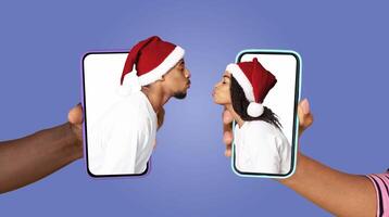 Two people in festive hats connect affectionately on their smartphones, sharing a kiss through the screens. They express love while embracing modern technology during the holiday season. photo