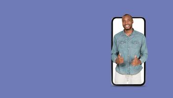 A young man smiles and gestures while holding a smartphone with a large screen. He seems to be using an application for social media or communication against a plain background. photo