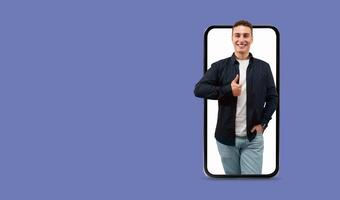 A young man stands confidently, smiling and giving a thumbs up gesture. He holds a smartphone with a large screen, symbolizing communication and connection in modern technology. photo