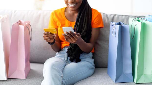 alegre afro mujer con crédito tarjeta y móvil teléfono sentado en sofá con regalo bolsas, confirmando Internet orden, utilizando en línea compras aplicación milenario negro dama adquisitivo en web foto