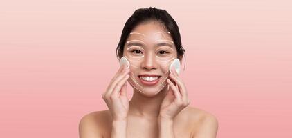 Asian woman is applying skin care products on her face, smiling as she enjoys her pampering routine. She is in a serene, isolated setting with a soft, minimalistic background. photo
