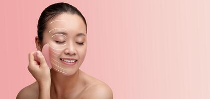 Asian woman is engaged in her skin care routine, gently applying a beauty tool to her cheek while smiling. The soft pink background adds to the serene atmosphere of her pampering session. photo