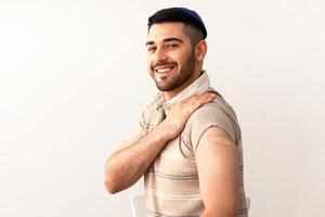 I Got My Coronavirus Vaccine. Happy Smiling Jewish Man In Kipa Showing Vaccinated Arm After Antiviral Injection For Covid Protection, Sitting On Chair Posing Over Studio Background, Looking At Camera photo
