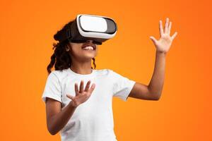 Excited young black girl in VR headset touching air during virtual reality experience at studio. Emotional kid exploring artificial computer world or playing game, isolated on yellow background photo