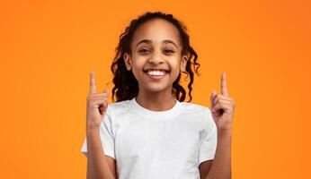 Look There. Portrait Of Smiling African American Girl Pointing With Two Fingers Up Above Her Head, Standing On Yellow Studio Background, Presenting Copy Space For Your Advertisement Or Information photo
