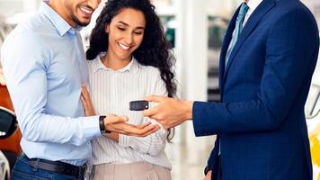 Smiling sales manager giving happy young middle eastern family key from brand new auto, side view. Emotional arabic couple buying car at luxury dealership salon. Car purchasing concept photo