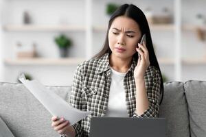 Irritated asian business lady working from home, sitting on couch with laptop, holding documents and having phone call, fighting with assistant or colleagues, copy space. Remote job concept photo