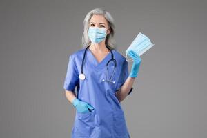 A middle aged woman in blue medical scrubs and a face mask stands confidently. She holds additional face masks in one hand, representing her role in healthcare. photo