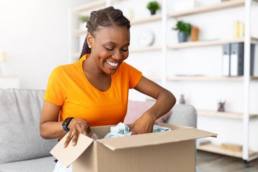 en línea entrega servicio. alegre africano americano mujer desempacar paquete o empaquetar caja con compra a hogar. contento hembra cliente satisfecho con ordenado bienes, mirando a nuevo ropa, compras en web foto