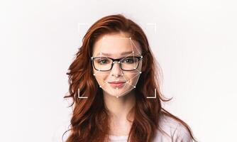 A woman with long red hair and glasses is the focus of facial recognition technology. The software outlines her facial features against a plain background, emphasizing identification capabilities. photo