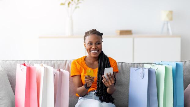 compras desde hogar. alegre negro mujer con crédito tarjeta y teléfono inteligente confirmando su en línea orden, sentado en sofá con regalo bolsas. joven africano americano dama adquisitivo artículos en Internet foto