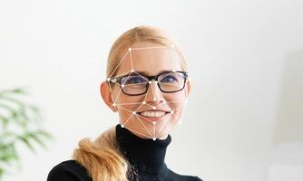 A person with glasses is smiling while facing the camera in a brightly lit modern office setting. The facial recognition technology overlays geometric points on their face. photo
