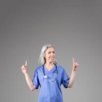 A middle aged woman wearing scrubs and a stethoscope smiles while pointing upwards at copy space. The setting is isolated with a plain gray background, emphasizing her enthusiasm and professionalism. photo