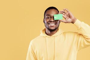 Positive Black Guy Posing With Credit Card Covering His Eye Standing Over Yellow Studio Background, Smiling To Camera. Finances, Bank Offer Concept. Panorama photo