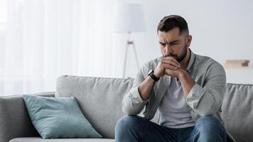 Thoughtful young serious handsome man sit on sofa at home, lost in thoughts, thinking about problem solving, feeling lonely, making important decision, having psychological problem in living room photo