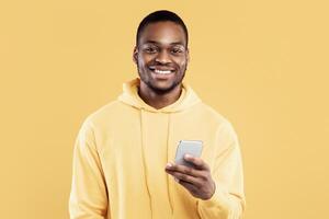 Happy Black Millennial Man Using Mobile Phone Smiling Looking At Camera Standing Over Yellow Studio Background. Guy Texting And Using New Application For Smartphone photo