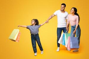 Seasonal Sales. Happy Middle Eastern Family With Little Daughter Carrying Bright Shopping Bags And Pointing Aside At Copy Space Over Yellow Background, Girl Showing Something To Parents, Panorama photo