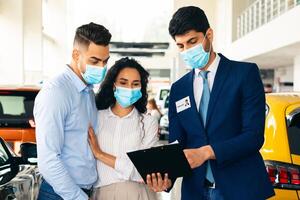 Millennial arab couple in protective face masks visiting auto showroom or exhibition while pandemic, muslim famile embracing while having conversation with salesman, looking at papers in his hands photo