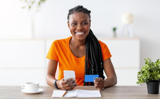 contento negro dama sentado a escritorio con Teléfono móvil y crédito tarjeta, compras en Internet a hogar. alegre africano americano mujer adquisitivo cosas en línea en web almacenar, sonriente a cámara adentro foto