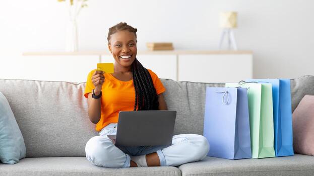 alegre negro mujer sentado en sofá con ordenador portátil computadora, participación crédito tarjeta, haciendo pago en en línea tienda desde hogar. contento cliente con regalo pantalones disfrutando electrónico Tienda servicio. comercio electrónico foto