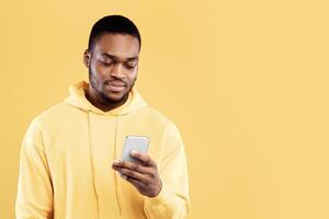 Calm African American Guy Using Smartphone Scrolling News And Texting Standing Over Yellow Studio Background. Millennial Man Using Mobile Application On His Phone. Technology And Gadgets photo