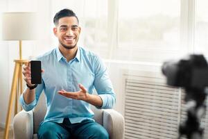 Arab guy influencer filming review of contemporary smartphone on dslr camera at home, mockup on screen. Joyful blogger advertising modern tech product, streaming live on web photo