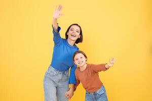 A mother and her preteen daughter are smiling and waving happily. They are holding hands, expressing affection and love in a cheerful setting with a vibrant yellow backdrop. photo