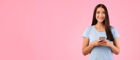 People And Technology Concept. Portrait of happy smiling young woman using smartphone standing isolated over yellow studio background, copy space. Lady chatting online, looking at camera photo