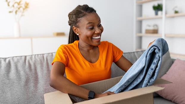 desempacar ropa. retrato de sonriente negro dama apertura cartulina caja, tomando fuera nuevo vaqueros, sentado en sofá a hogar. joven africano americano mujer satisfecho con en línea compras y entrega Servicio foto