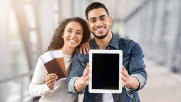 Arab Couple Demonstrating Digital Tablet With Black Screen While Standing In Airport With Passports And Tickets In Hands, Young Spouses Showing Copy Space For Your Design Or Advertisement, Mockup photo