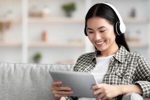Pretty asian lady watching movie, using wireless headset and pad while resting on couch at home, copy space. Happy young woman enjoying new entertaining mobile application on digital tablet photo