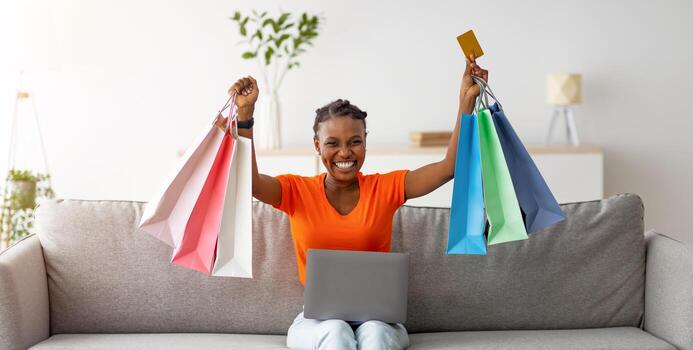 emocionado afro dama compras en línea, sentado en sofá con ordenador portátil y crédito tarjeta, levantamiento regalo pantalones encima su cabeza a hogar. milenario negro mujer adquisitivo en web, comprando en descuento adentro foto