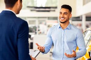 Rich handsome middle-eastern young man visiting newest auto showroom or dealership salon, buying auto, talking to sales manager and smiling, asking for help with choosing nice automobile photo