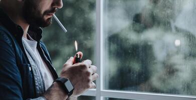 Suffering some problem, feeling desperate, crisis and depression. Habit and addiction. Sad millennial bearded guy lights cigarette with lighter, on window background, cropped, panorama, copy space photo