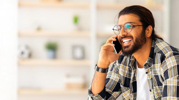 alegre árabe chico con largo pelo y lentes relajante en sofá a hogar, teniendo teléfono hablar con amigos, mirando a Copiar espacio y sonriente, vistiendo casual atuendo, descansando a fin de semana foto