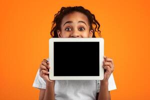 Great Application. Excited Black Girl Holding And Showing Digital Tablet With Empty Screen For Mockup, Covering Face And Mouth, Recommending New App Posing Standing Over Yellow Studio Background photo