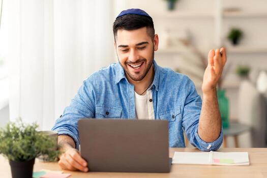 distante comunicación concepto. retrato de contento judío masculino en kipá y casual camisa haciendo llamada sentado a escritorio a hogar oficina y hablando con colegas, estudiantes o familia, explicando foto