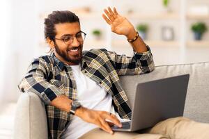 Cheerful long-haired, bearded indian guy sitting on couch in living room, using laptop, having conference with client or girlfriend from home, smiling and waving at notebook screen, copy space photo