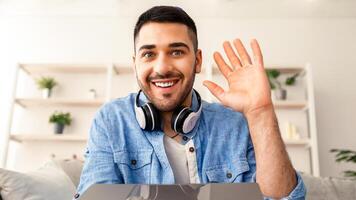 Greeting Concept. Portrait of excited young man waving hello with hand looking at camera, using laptop at home office. Cheerful guy saying hi or goodbye during call, webcam view photo