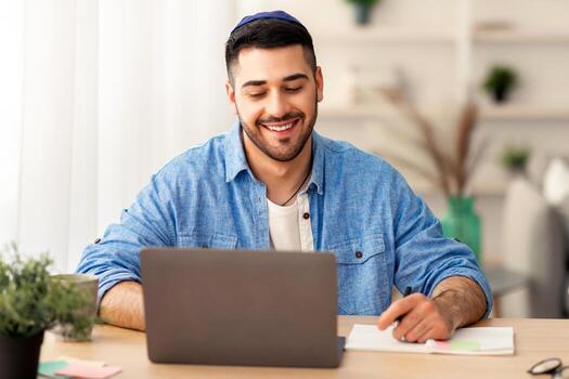 retrato de contento israelí hombre vistiendo kipah trabajando en ordenador portátil sentado a lugar de trabajo en moderno hogar oficina. negocio y emprendimiento, exitoso carrera y distancia trabajo concepto. selectivo atención foto