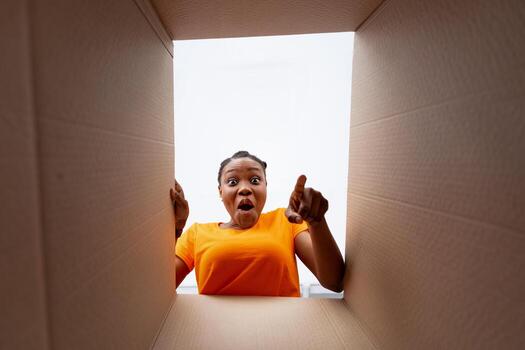 emocionado africano americano dama señalando dentro paquete o empaquetar caja, recepción en línea orden, fondo vista. satisfecho negro mujer disfrutando entregado bienes desde web Tienda durante codicioso encierro foto