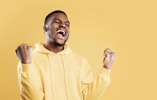 Emotional Black Man Shouting Shaking Fists With Eyes Closed Celebrating Big Success Over Yellow Studio Background. Celebration Of Victory, Joy Emotion Concept. Panorama With Free Space For Text photo