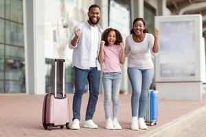Holiday Trip After Lockdown. Full body length portrait of excited African American mum, daddy and daughter going on a journey. Family shaking clenched fists, making win gesture, standing near airport photo