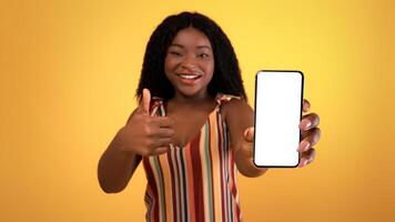 Lovely black woman demonstrating cellphone with empty black screen, showing thumb up, recommending new cool mobile shopping app on orange studio background, mockup for design photo