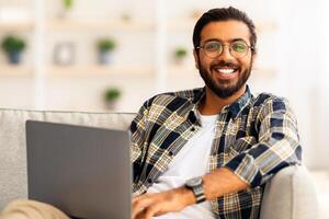Positive indian guy reclining on couch with laptop, watching movie or chatting with ladies on dating website while relaxing at weekend, middle-eastern freelancer working from home, copy space photo