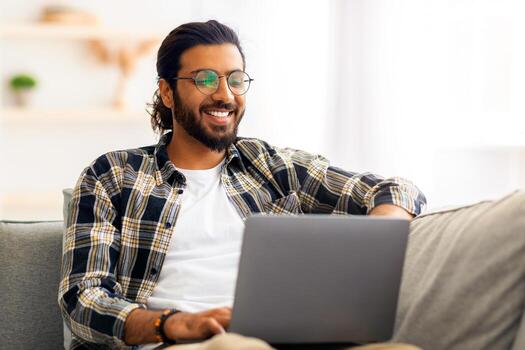 contento Medio este joven hombre en casual persona de libre dedicación disfrutando su en línea trabajo, reclinable en sofá a hogar, utilizando moderno computadora portátil, Copiar espacio. alegre indio chico websurf o compras en línea foto