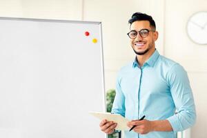 Joyful guy English teacher explaining foreign language rules near blackboard, standing with clipboard, smiling at camera in office. Young Arab tutor teaching online course, giving lecture or webinar photo